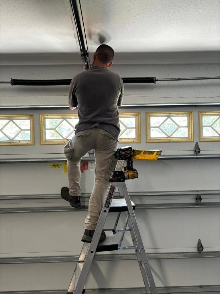 Technician on a stepladder reaching the torsion shaft above a windowed garage door