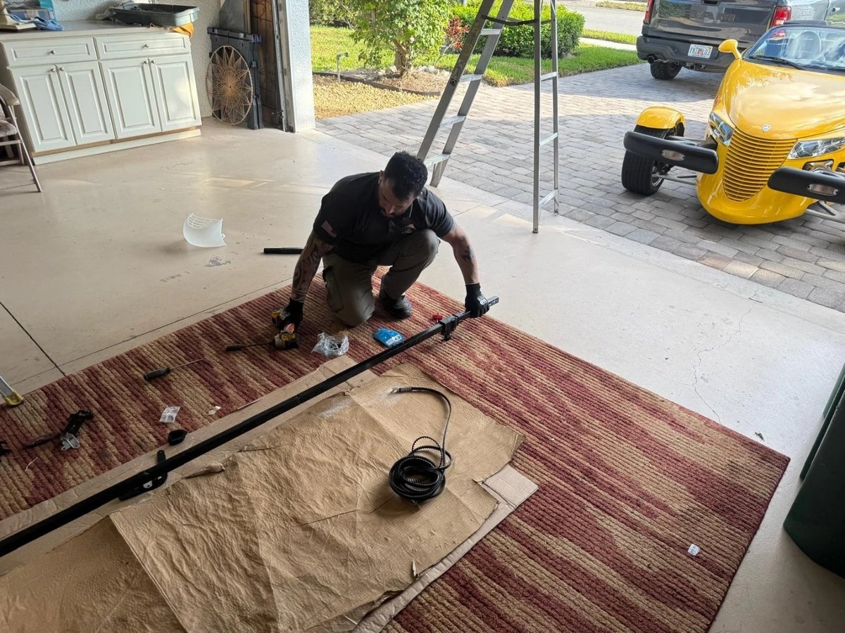 Technician kneeling on a rug beside a long metal torsion rod laid out on a garage floor