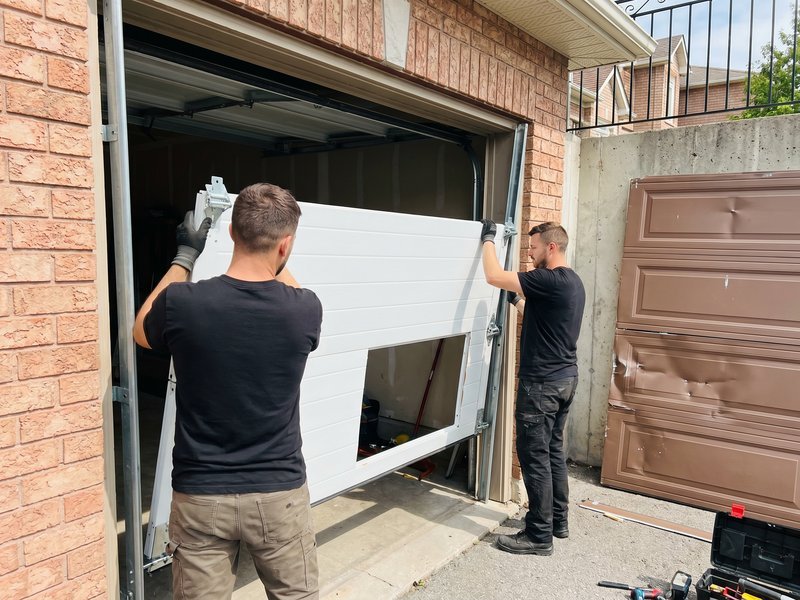 Two people lifting a white replacement panel into a garage door opening on a brick-front house