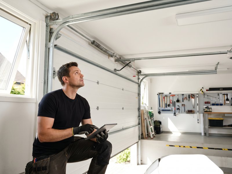 Technician in a black shirt kneeling beside a closed paneled garage door with a tool in hand