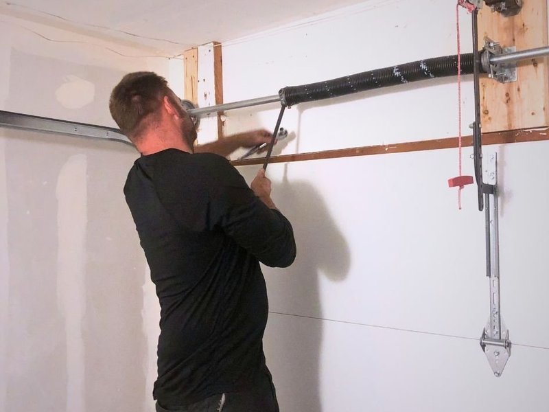 Technician reaching up to the spring and lift cable hardware beside a garage door